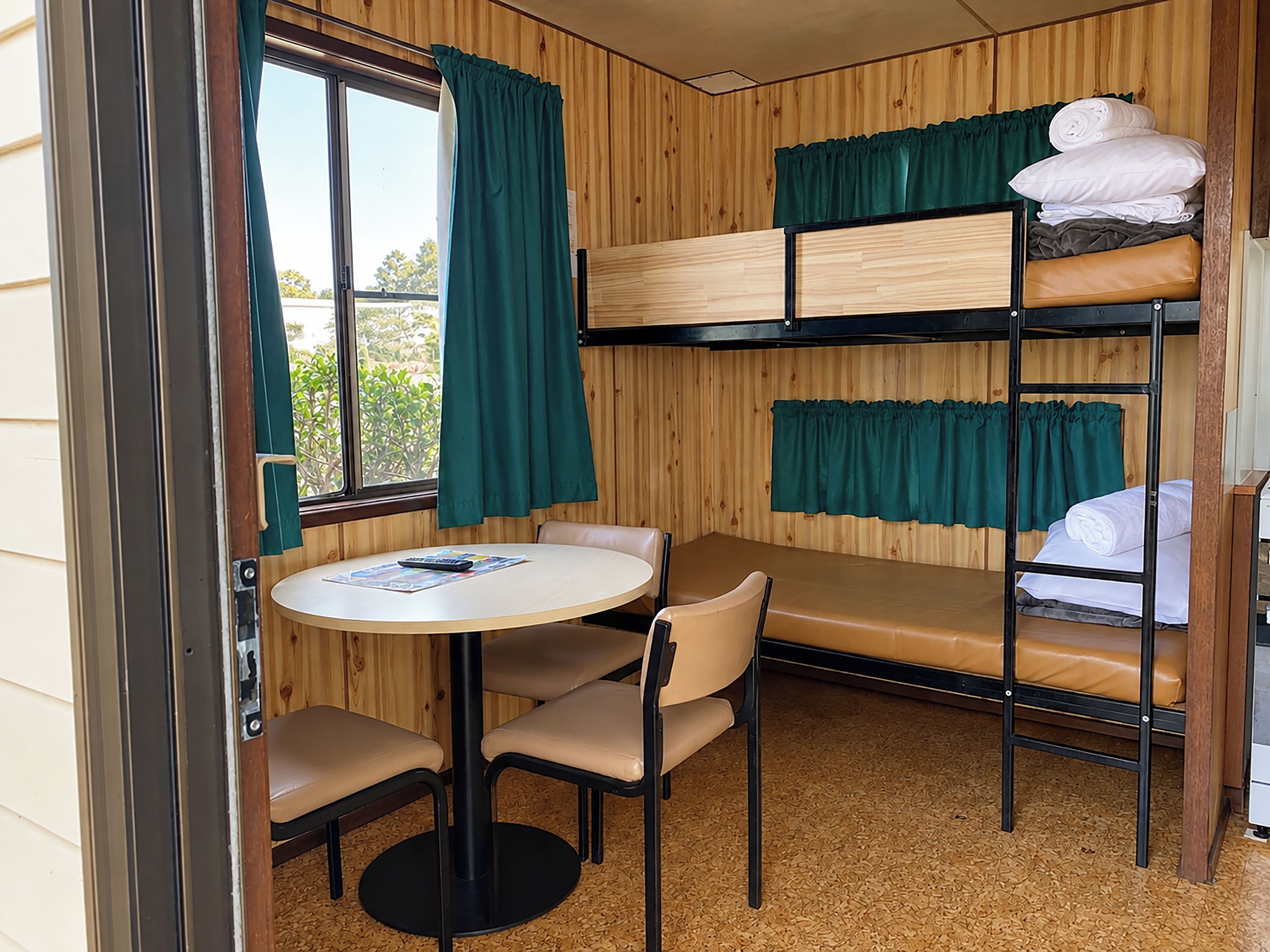 A bunk bed room with a table and chairs, a window with curtains, and a view of greenery outside.
