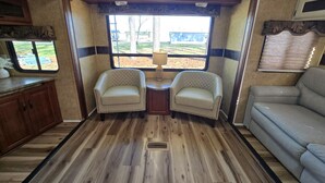 Ein kompaktes Wohnmobil-Interieur mit zwei gepolsterten Sesseln, einer Sitzecke, Holzboden und einem Fenster mit Blick ins Freie.