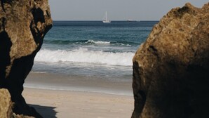 Una playa con un velero a lo lejos, enmarcada por afloramientos rocosos.