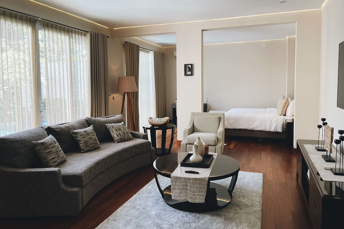 Habitación de hotel moderna con sofá, sillón y cama. Se observa una lámpara, una mesita con un florero y un cuadro en la pared.