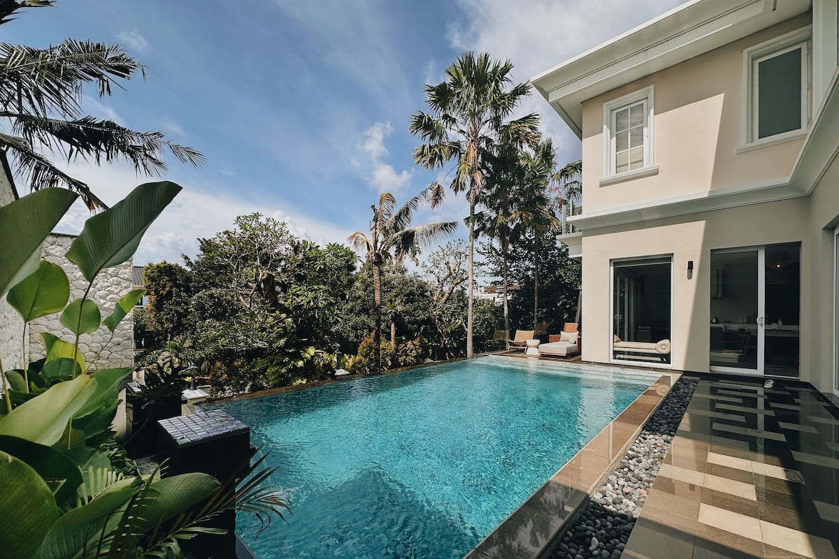 Un área de piscina exterior moderna con una piscina de color azul cristalino, rodeada de exuberante vegetación y un muro de piedra.