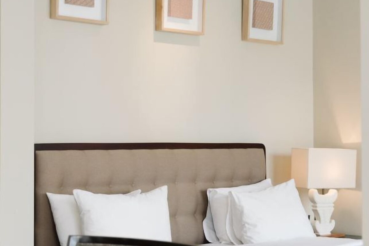 A hotel room with a bed, white pillows, a headboard, a nightstand with a lamp, and framed pictures on the wall.