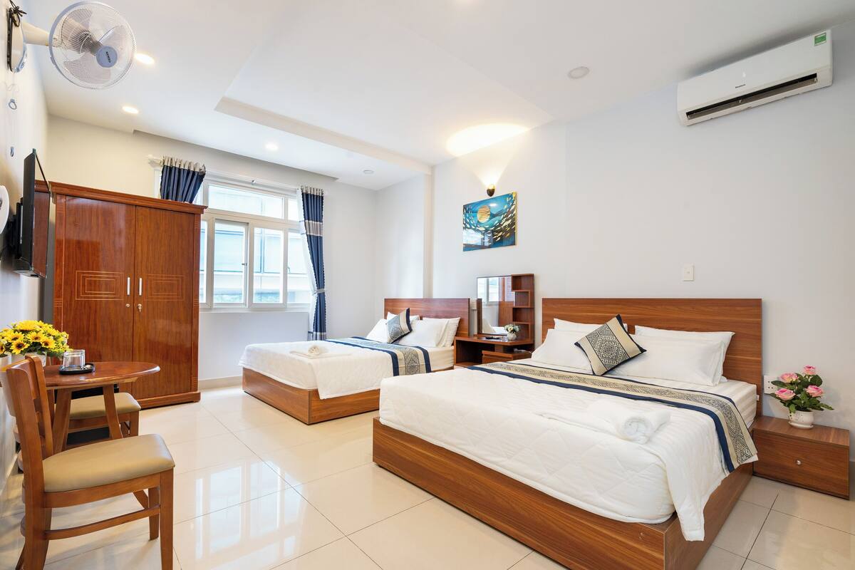 A hotel room with two beds, a wooden wardrobe, a desk with a chair, and a dining table with flowers.