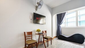 A room with a TV mounted on the wall, two wooden chairs, a small round table with a vase of flowers, a standing fan, and a guitar case on the floor. - Green Airport Hotel (Ho Chi Minh City)