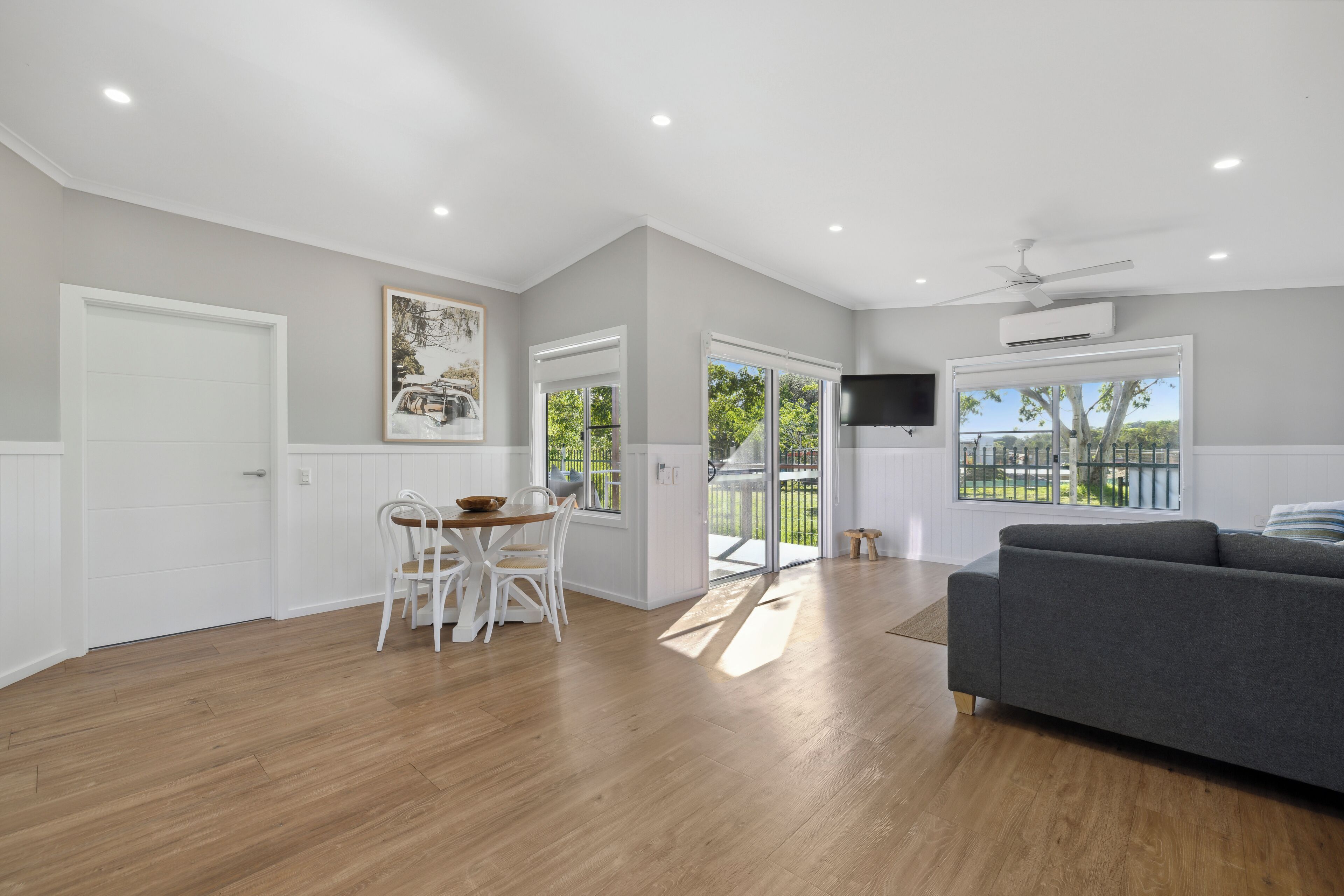 A spacious living area with a grey sofa, wooden flooring, a dining table with chairs, and a TV mounted on the wall.