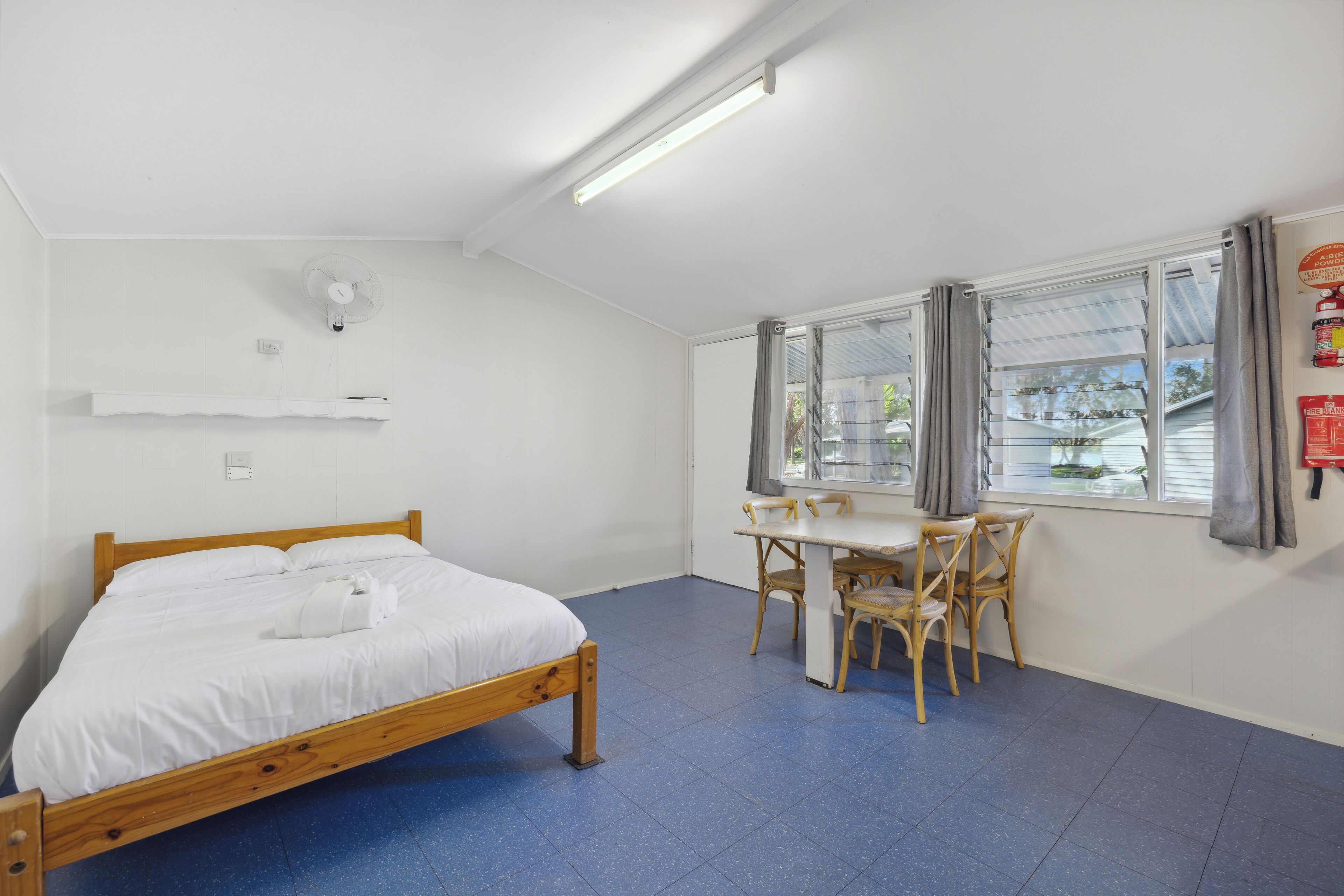 A room with a bed, a desk, and chairs. There is a window with curtains, a ceiling fan, and a fire extinguisher on the wall.