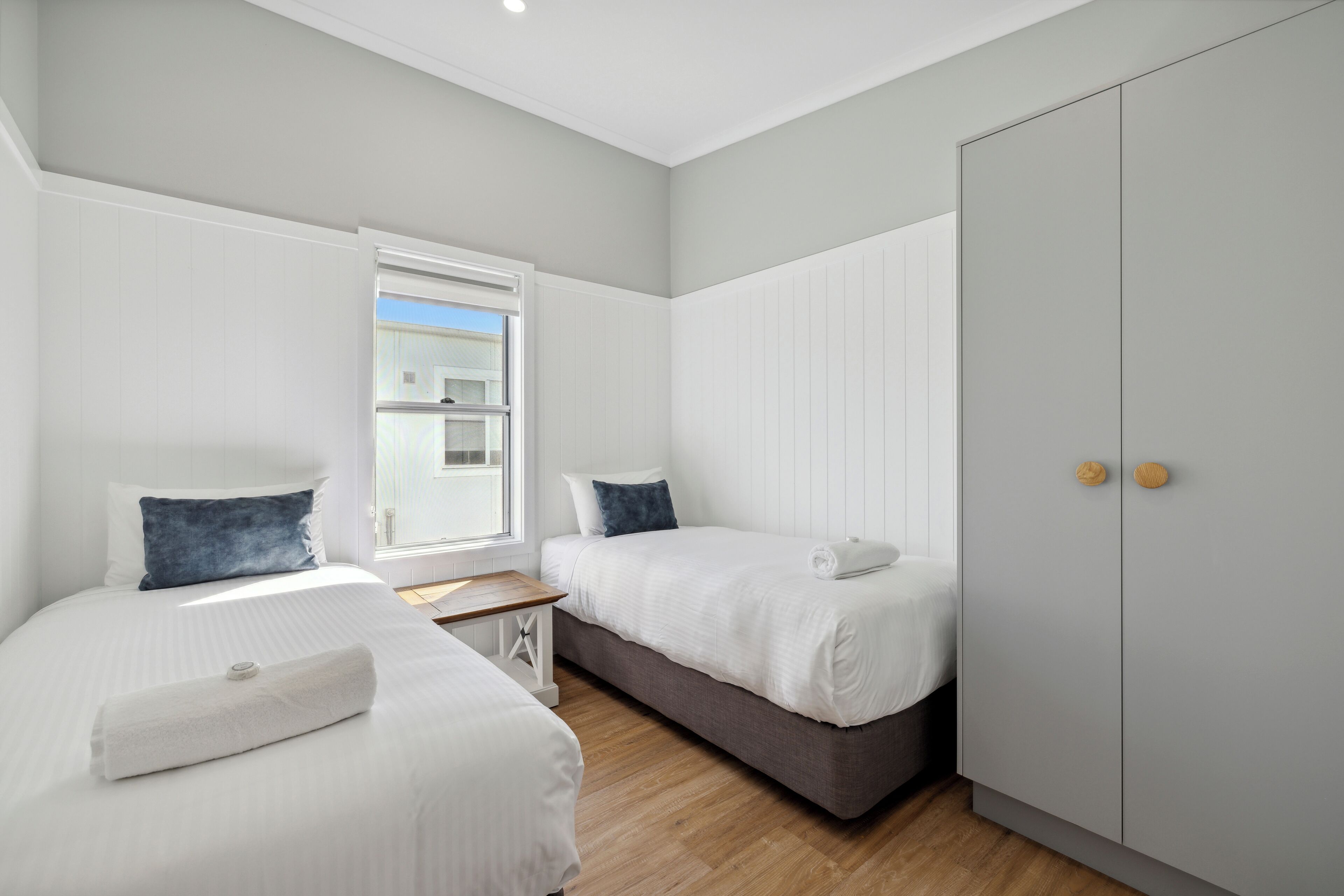 A modern hotel room with two single beds, a large wardrobe, and a small table.