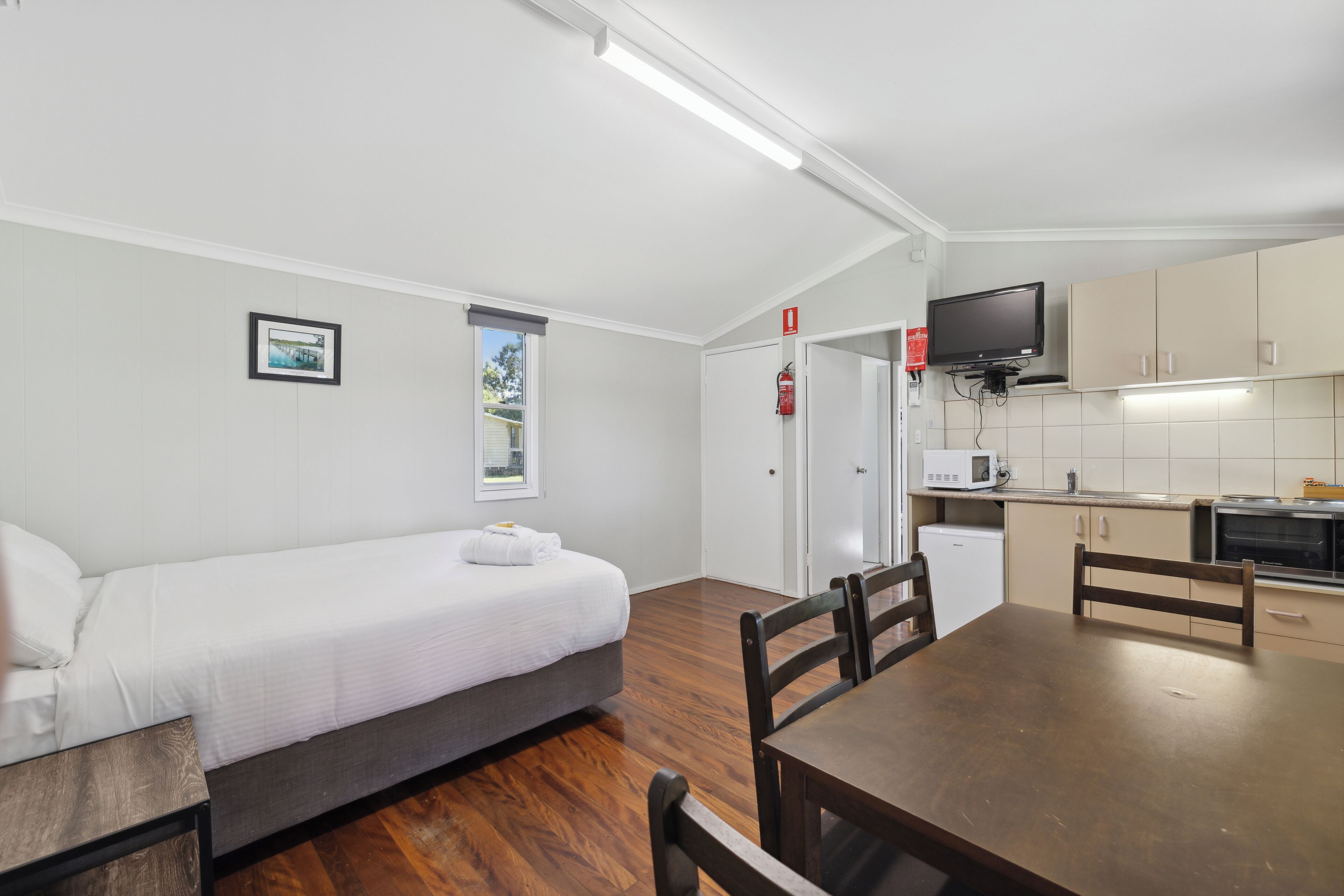 A room with a bed, a dining table, chairs, a kitchen area with a microwave, and a television.