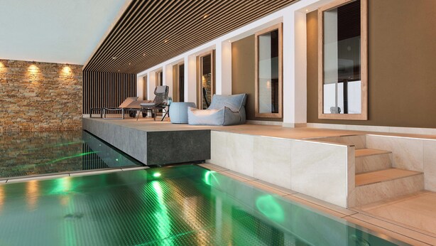 Un'area piscina interna moderna con parete in pietra, soffitto in listelli di legno e un angolo relax con tavolo e sedie.