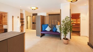 Una moderna hall d'hotel con una reception, un'area salotto con divano e cuscini, un pannello divisorio in legno e una pianta in vaso.