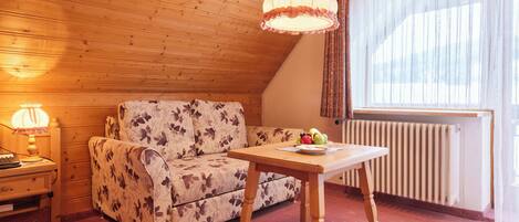 Una habitación acogedora con techo de madera, un sofá floral, una mesa de madera con fruta, una lámpara y un radiador.