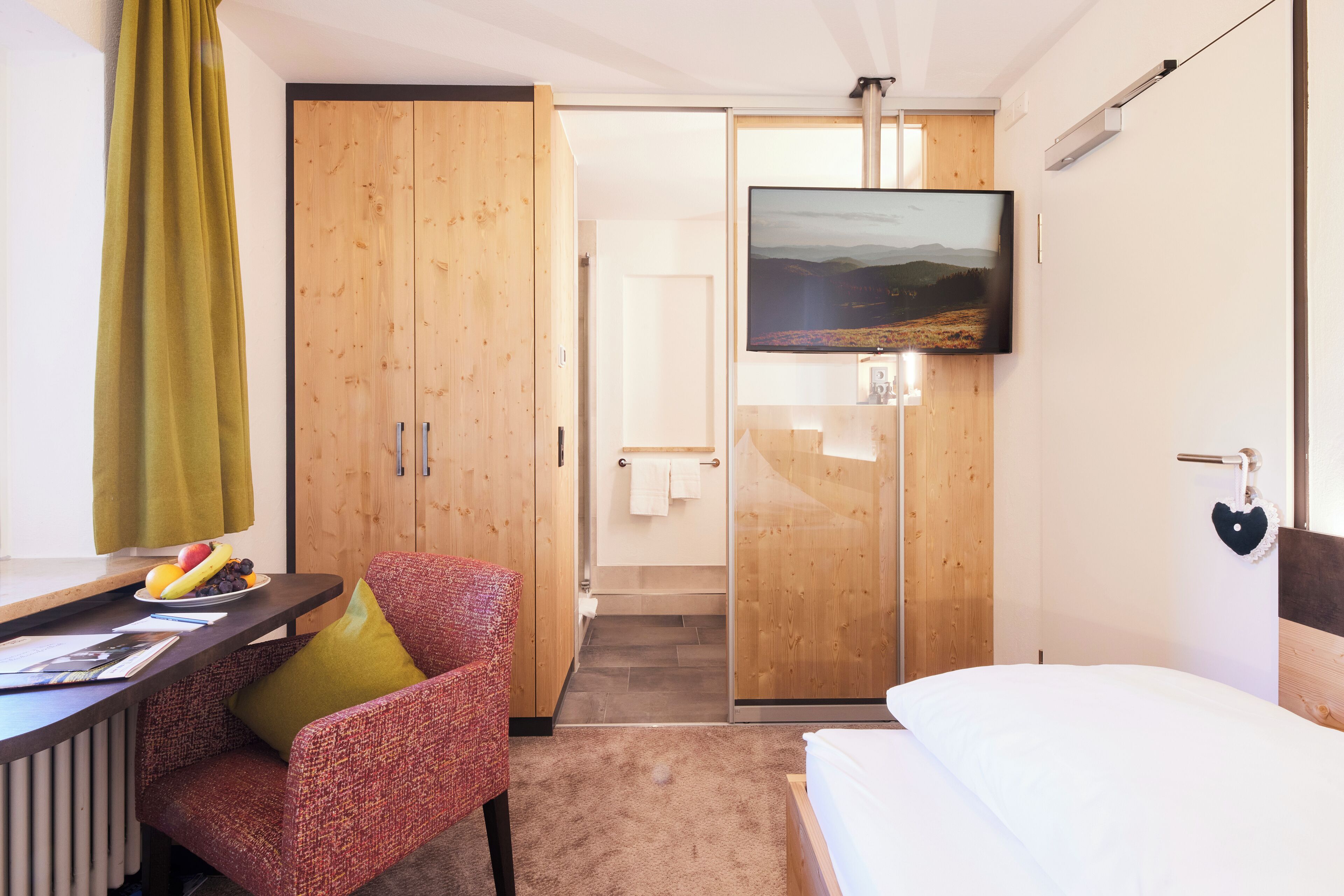 Una habitación de hotel con una cama, un escritorio con una silla, un televisor montado en la pared y un armario.