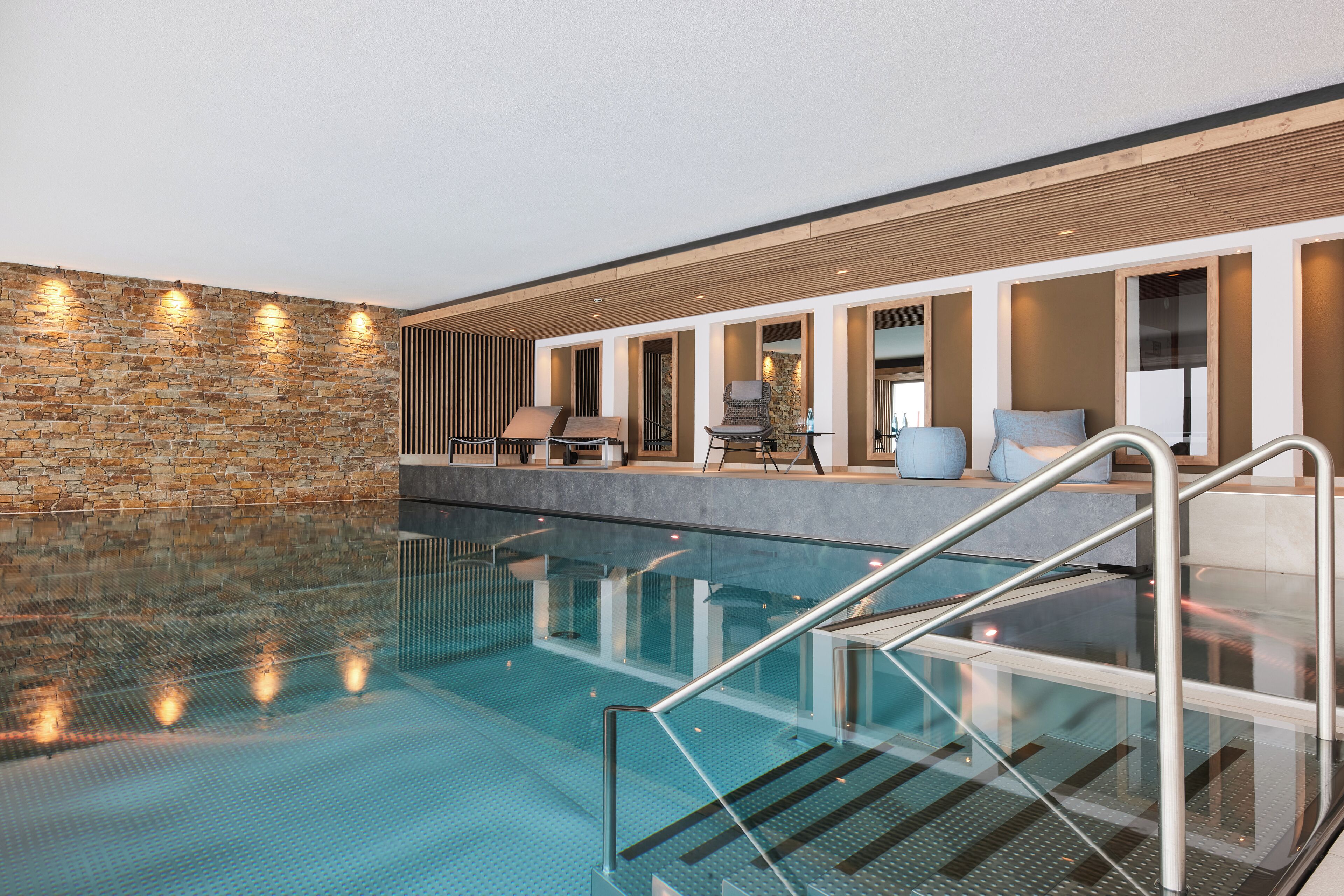 An indoor swimming pool with clear glass walls, a stone wall on one side, and a seating area with chairs and a table.