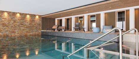 An indoor swimming pool with clear glass walls, a stone wall on one side, and a seating area with chairs and a table.