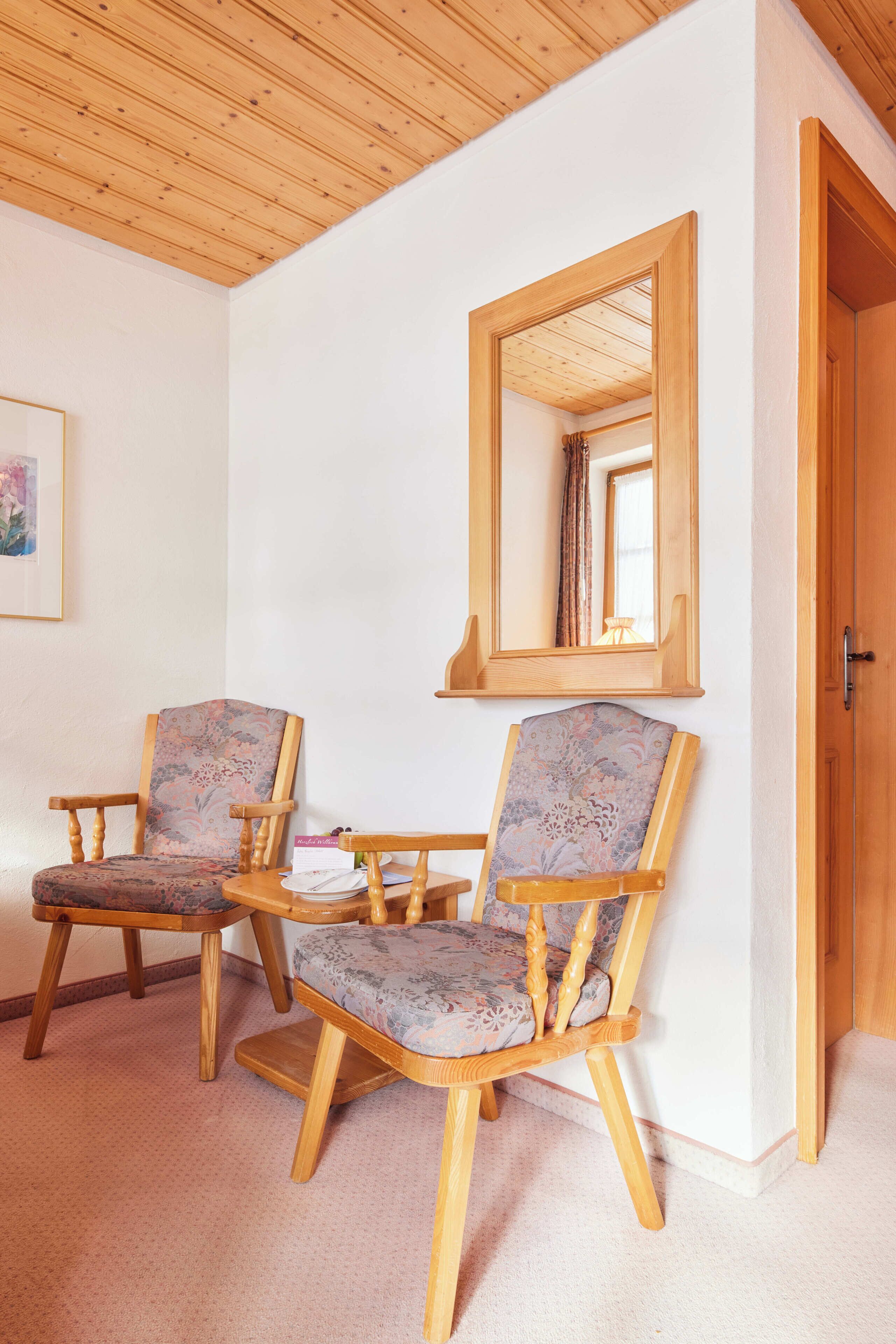 A room with two wooden chairs with floral upholstery, a small table with a plate and a vase, and a mirror on the wall.