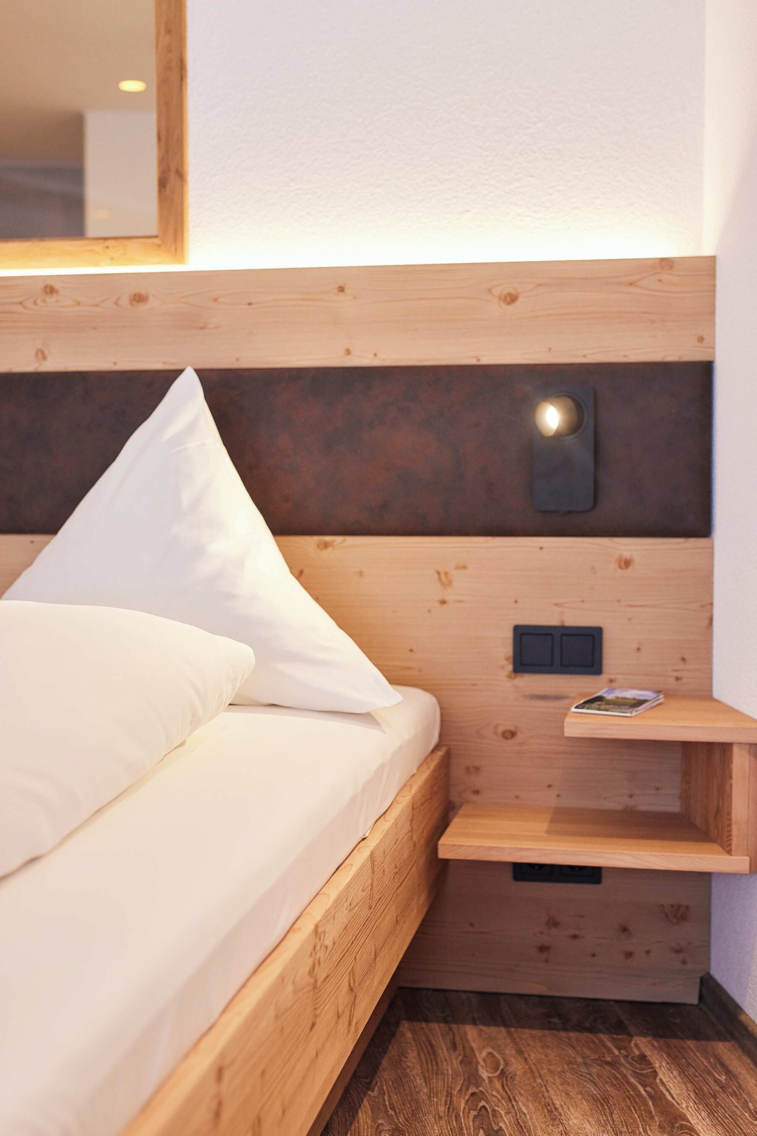 Ein ordentlich gemachtes Bett mit weißen Leinen, ein hölzernes Kopfende, ein kleiner Nachttisch mit einer Zeitschrift und eine Wandleuchte.