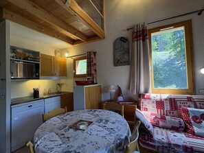 Une pièce chaleureuse avec une table ronde, un canapé, un coin cuisine doté de meubles en bois et une grande fenêtre donnant sur les arbres.