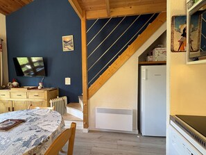 Une pièce avec un escalier en bois, un réfrigérateur blanc, une commode en bois, une table recouverte d’une nappe à motifs et un téléviseur fixé au mur.