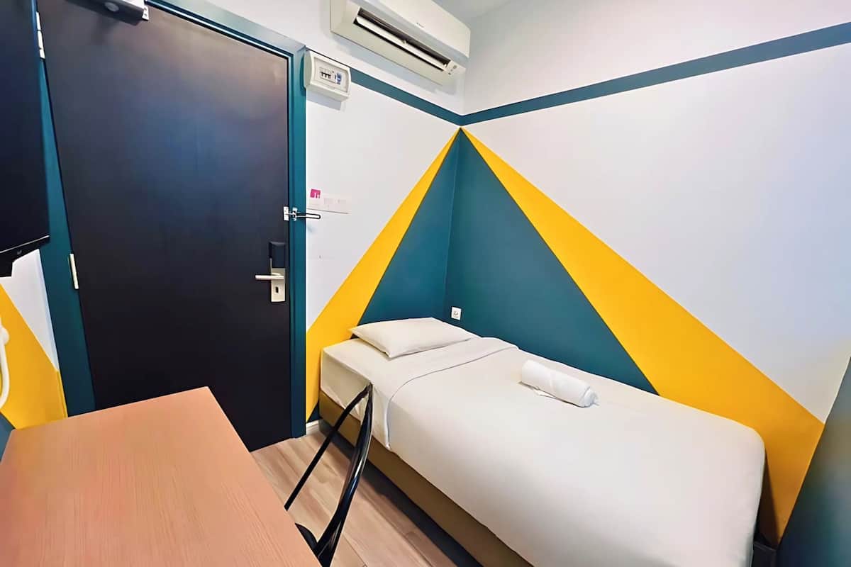 Quarto pequeno e bem arrumado, com cama, escrivaninha e ar condicionado na parede.
