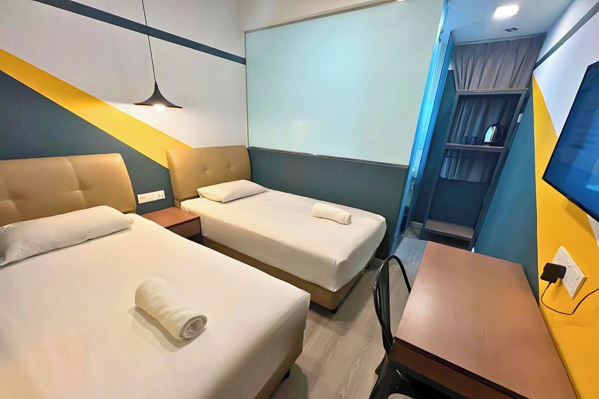Quarto de hotel com duas camas, uma escrivaninha, uma cadeira, uma televisão e uma janela com cortinas.