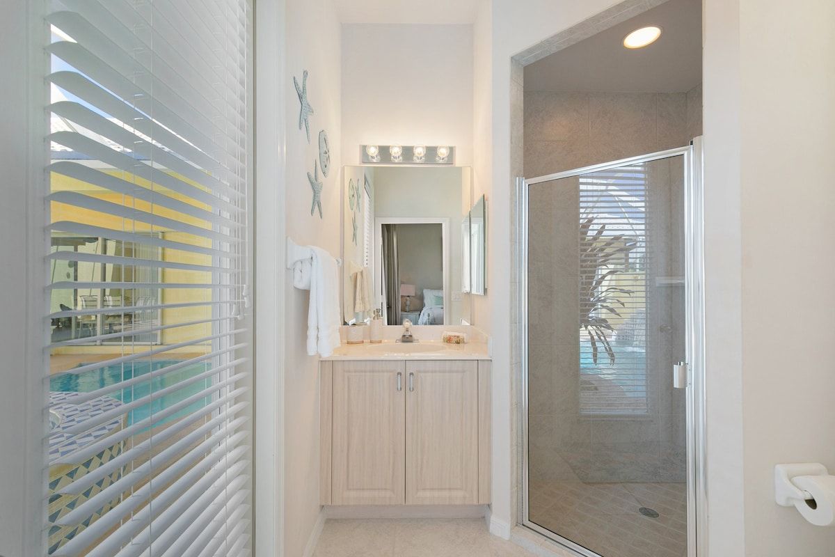 A bathroom with a shower, a vanity with a mirror, and a window with blinds.