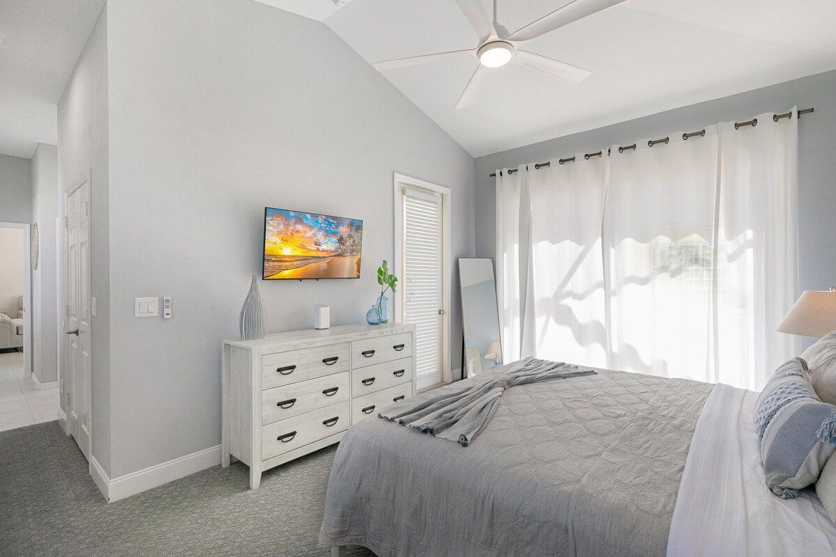 Un dormitorio con cama, cómoda, cuadro, ventilador de techo y ventana con cortinas.