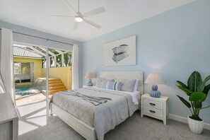 A bedroom with a bed, bedside tables, a ceiling fan, a painting, and a view of a pool and outdoor area.