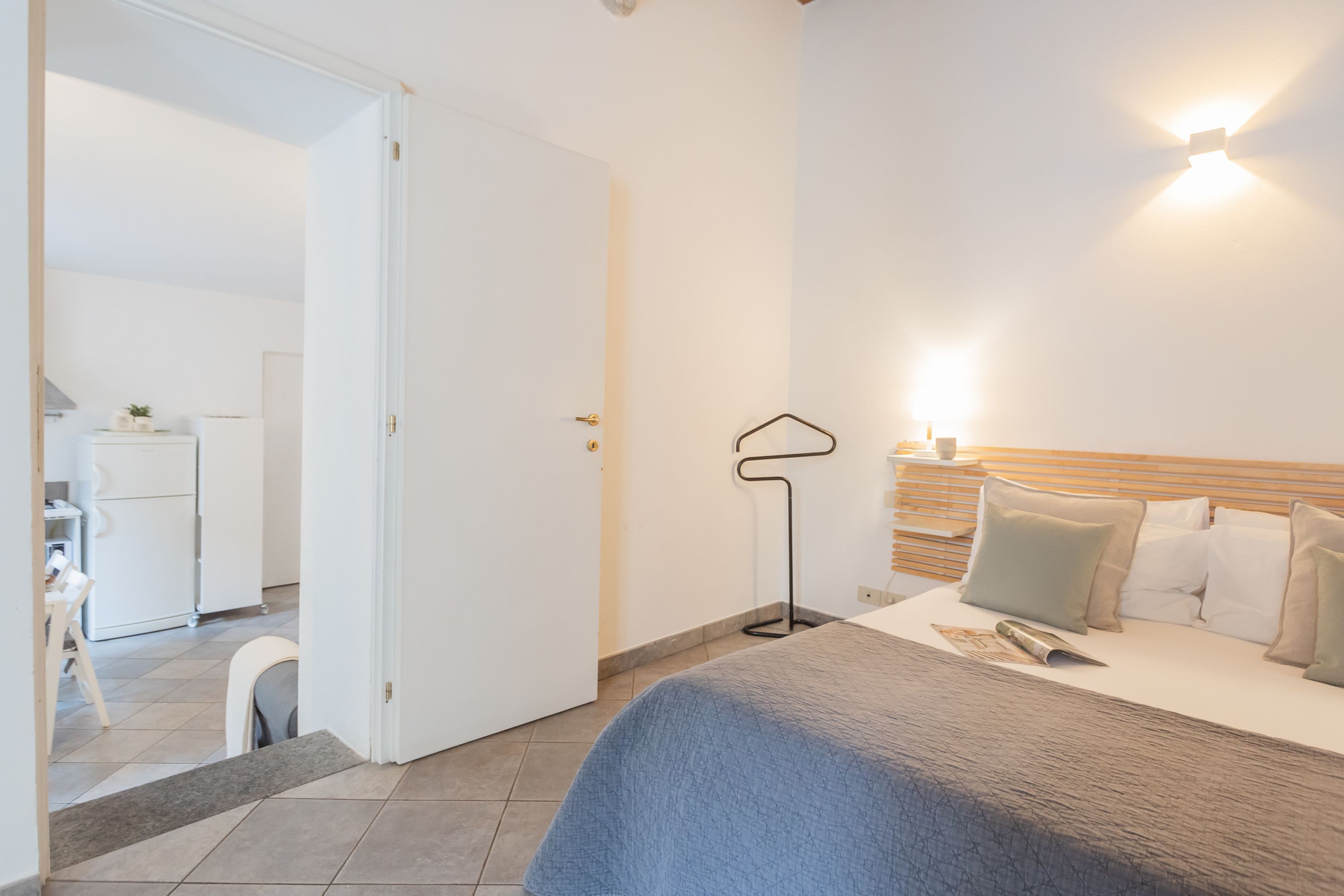 Une chambre à coucher comprenant un lit, une table de chevet, une lampe et donnant sur la cuisine et l’espace repas.