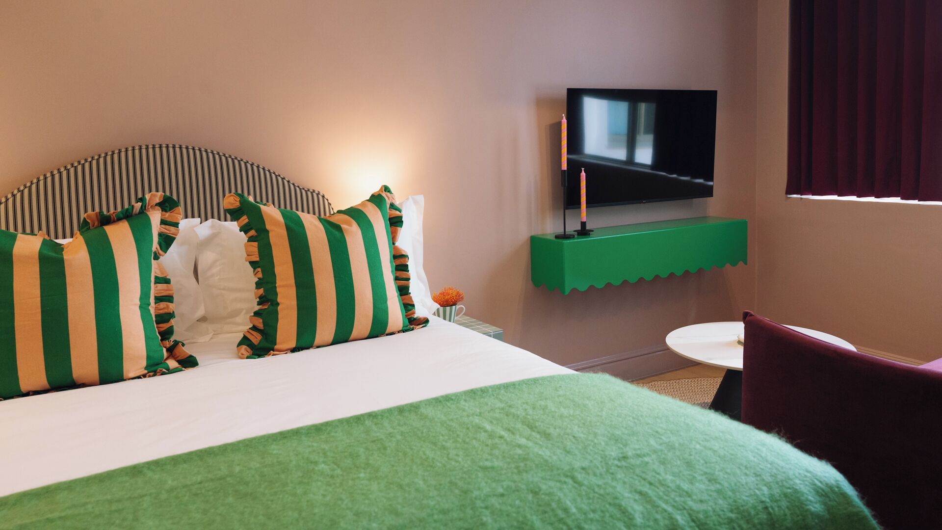 A hotel room with a bed, a TV mounted on a green shelf, a small table, and a chair.