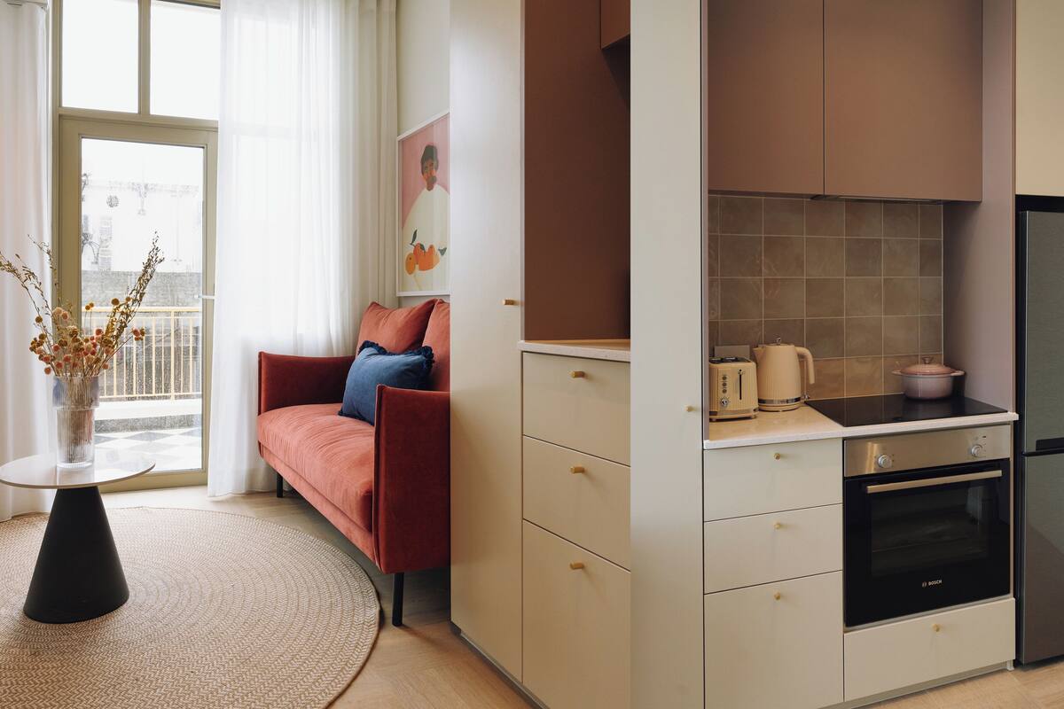 Een compacte keuken met een moderne Bosch oven, een klein zitehoek met een bank en een vaas gedroogde bloemen, en uitzicht op de stad door een groot raam.