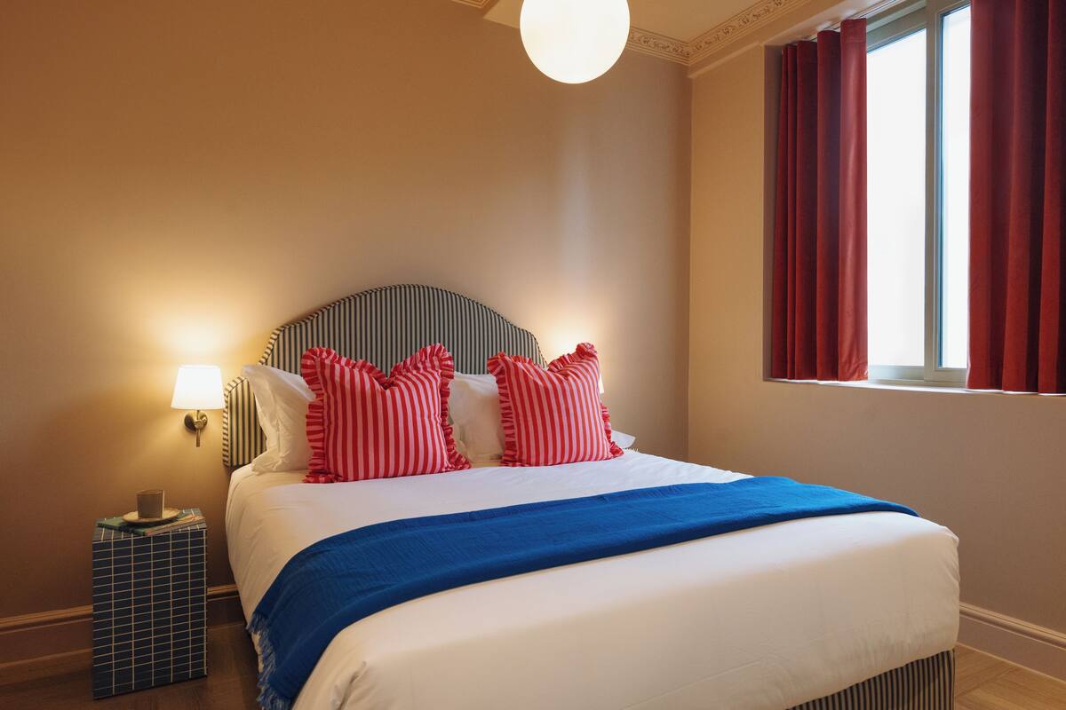 Een netjes opgemaakt bed met een gestreept hoofdbord, een blauwe plaid en rode kussens. Er staat een bedlamp op een klein tafeltje naast het bed.