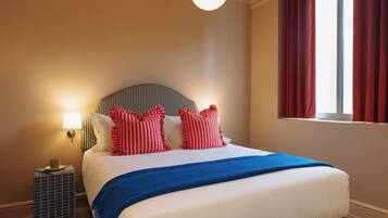 A neatly made bed with a striped headboard, a blue throw blanket, and red pillows. There is a bedside lamp on a small table beside the bed.