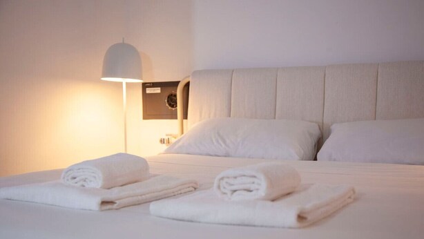 A neatly made bed with two rolled towels, a wall-mounted lamp, and a nightstand with a clock. - Navigli Beautiful Apartment in MILAN Near TRAM (Milano)