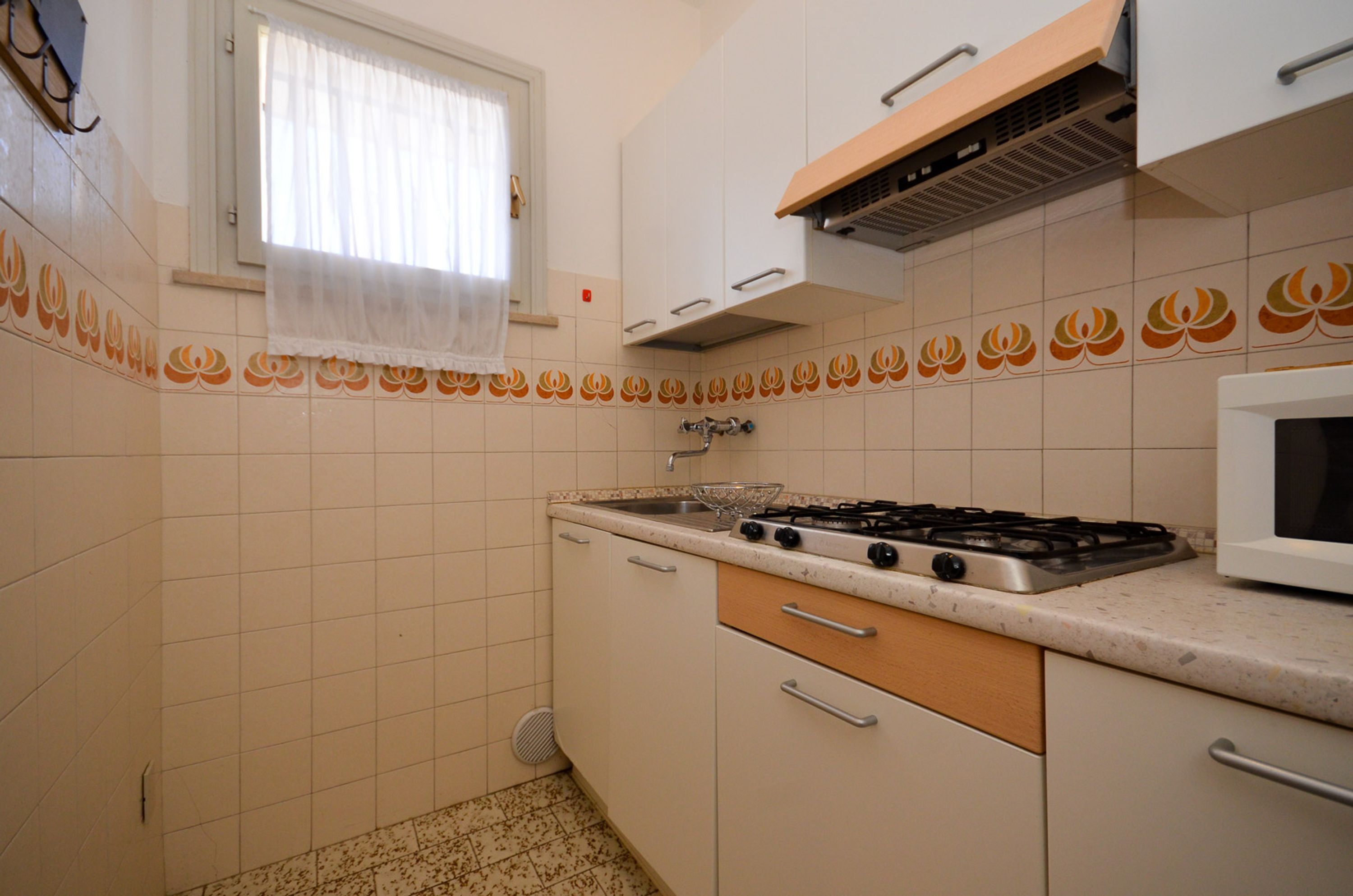 Una cocina con armarios blancos, estufa a gas, microondas y una ventana con cortina blanca.