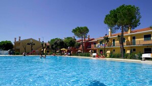 A swimming pool with people in the water, surrounded by yellow buildings with balconies and trees. - Michelangelo C-6 in Bibione (San Michele al Tagliamento)