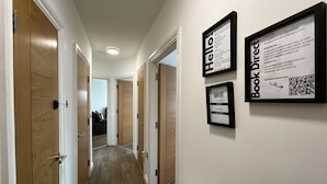 A hallway with wooden doors, a framed 'Hello' sign, a 'Book Direct' sign, and a framed WiFi password sign. - City Retreat Birmingham - Modern 2 Bed Apartment (Birmingham)