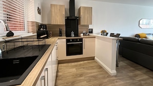 A modern kitchen with wooden cabinets, a black range hood, and a dining area with a black sofa and a small table. - City Retreat Birmingham - Modern 2 Bed Apartment (Birmingham)
