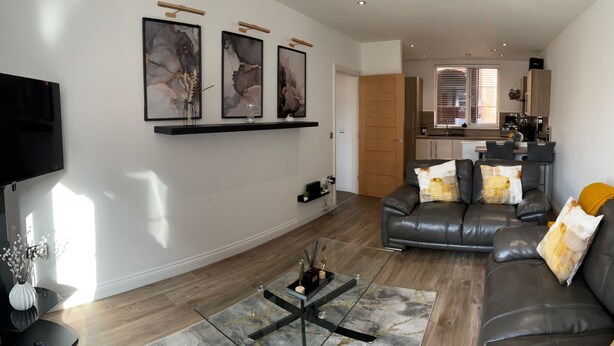 A modern living room with a leather sofa, a coffee table, and a TV. There is a kitchen in the background with a dining area. - City Retreat Birmingham - Modern 2 Bed Apartment (Birmingham)