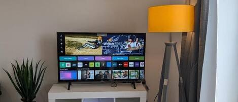 Una sala de estar con un televisor de pantalla plana sobre un soporte blanco, una planta en maceta, una lámpara de pie amarilla y un cuadro de un elefante en la pared.