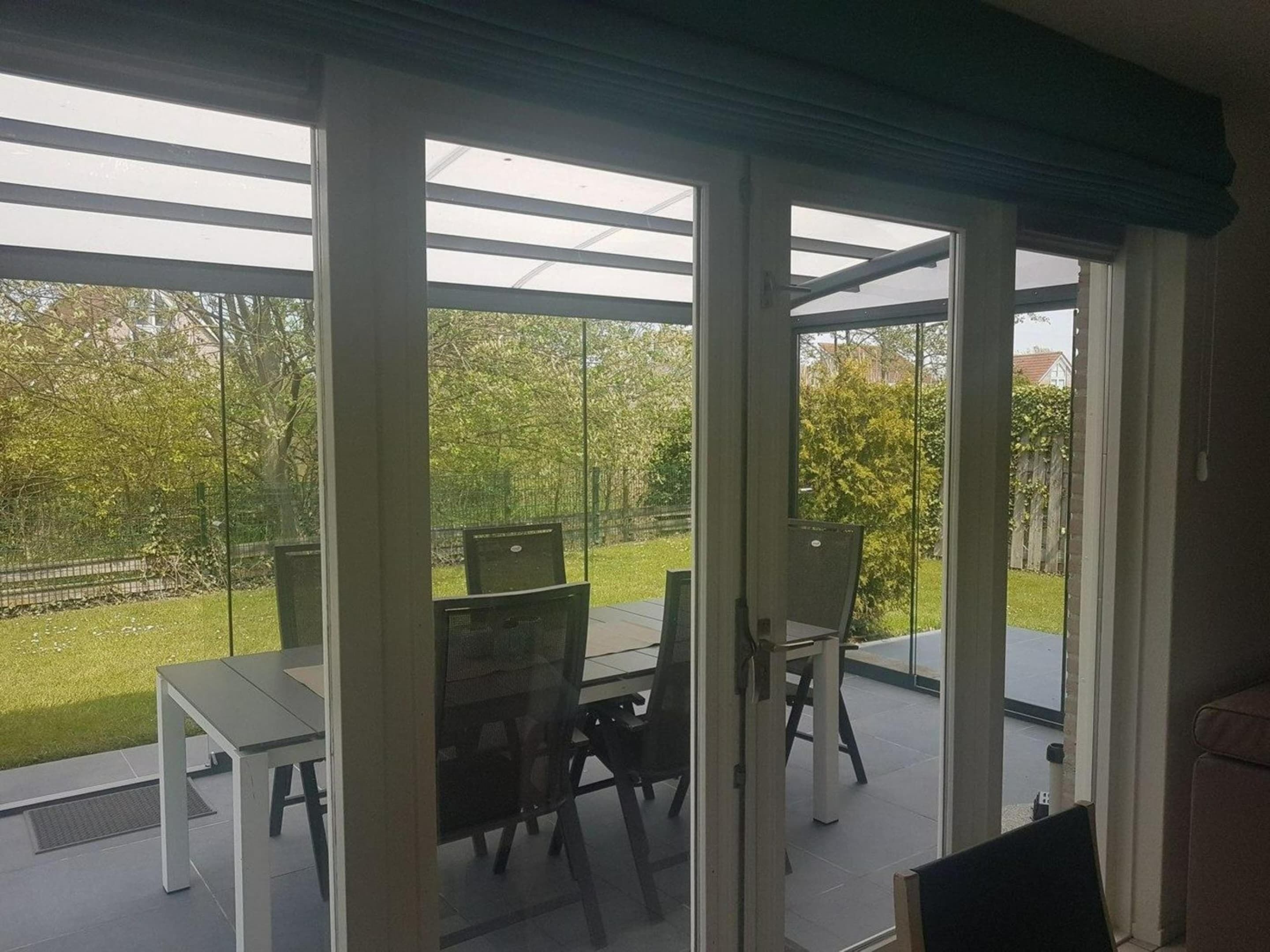 A covered patio with a table and chairs, large windows, and a view of a garden.