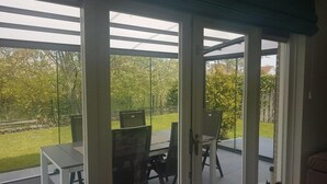 A covered patio with a table and chairs, large windows, and a view of a garden. - Bungalow in Breskens Near Beach & Lighthouse (Breskens)