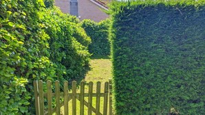 A wooden gate leading into a garden with a hedge on one side and a building in the background. - Bungalow in Breskens Near Beach & Lighthouse (Breskens)