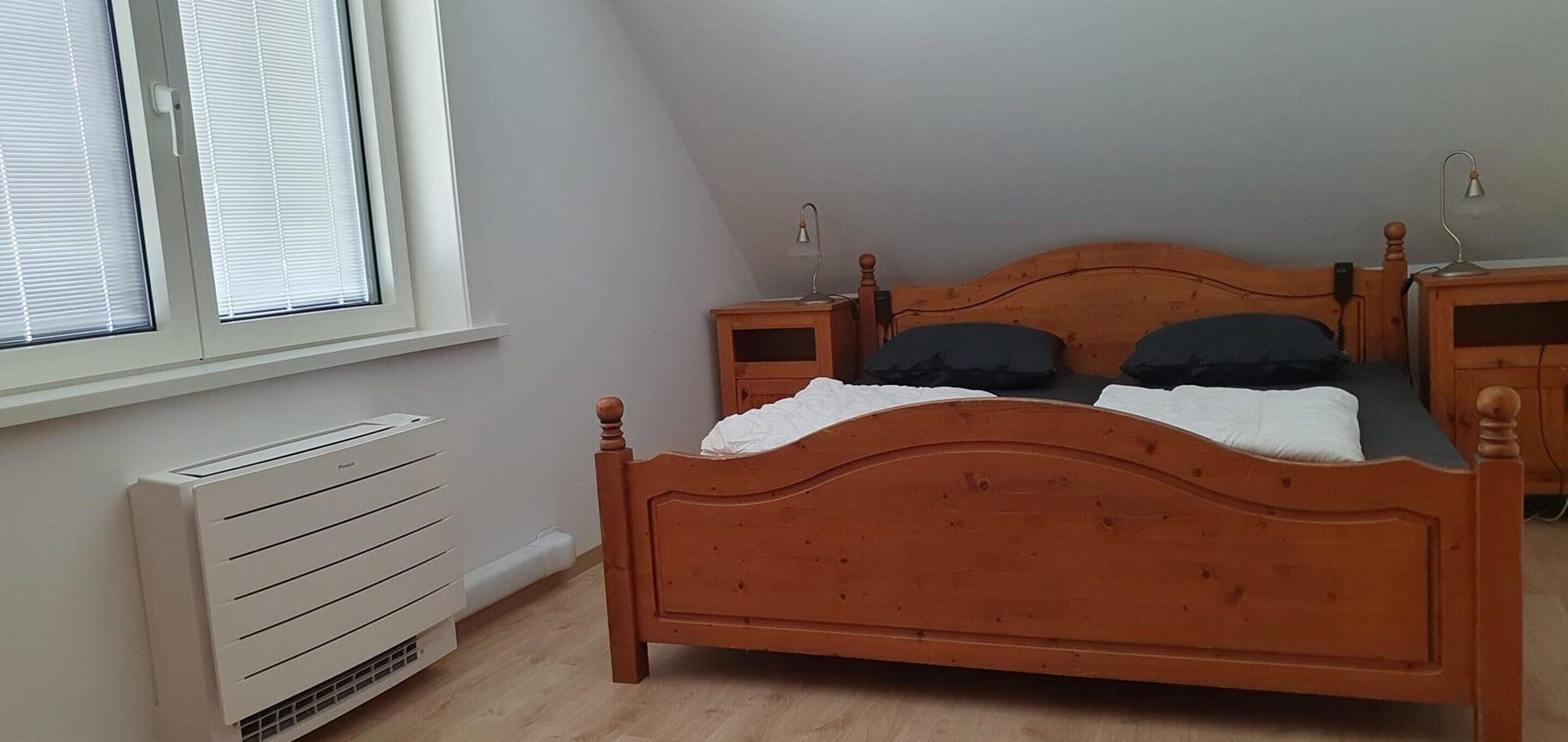 A bedroom with a wooden bed, two bedside tables, a nightstand, a window with blinds, and a wall-mounted heater.