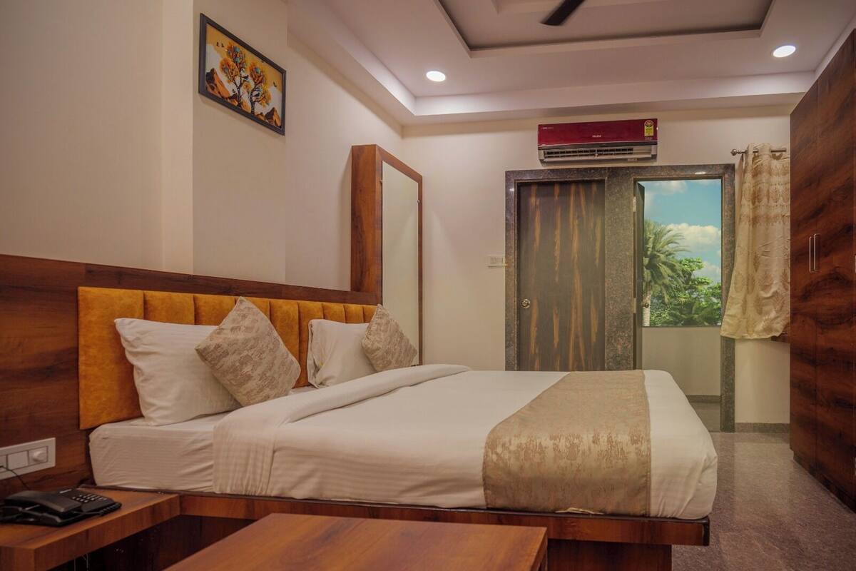 A hotel room with a wooden headboard, a bed with white linens, a bedside table with a phone, and a view of greenery through a door.