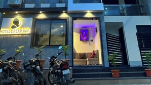 A modern hotel with a sign reading "HOTEL DREAM INN." It features multiple balconies with potted plants, a brightly lit entrance, and several motorcycles parked outside. - Hotel Dream Inn (Sanwer)