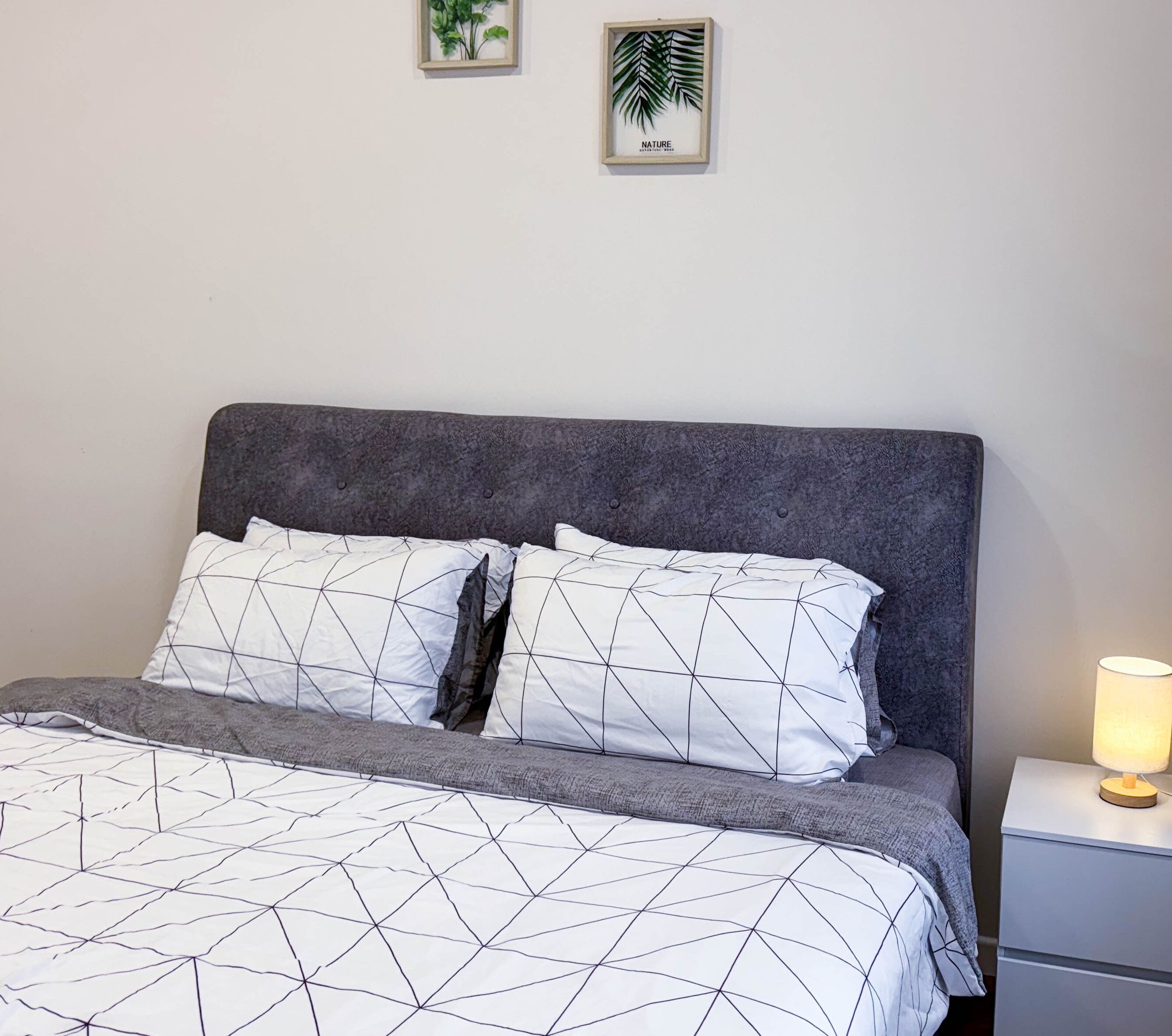 Un lit bien fait, avec une couvre-lit à motifs géométriques et des oreillers, une tête de lit rembourrée de couleur grise et une table de chevet avec une lampe. Deux tableaux encadrés représentant des paysages sont accrochés au mur au-dessus du lit.