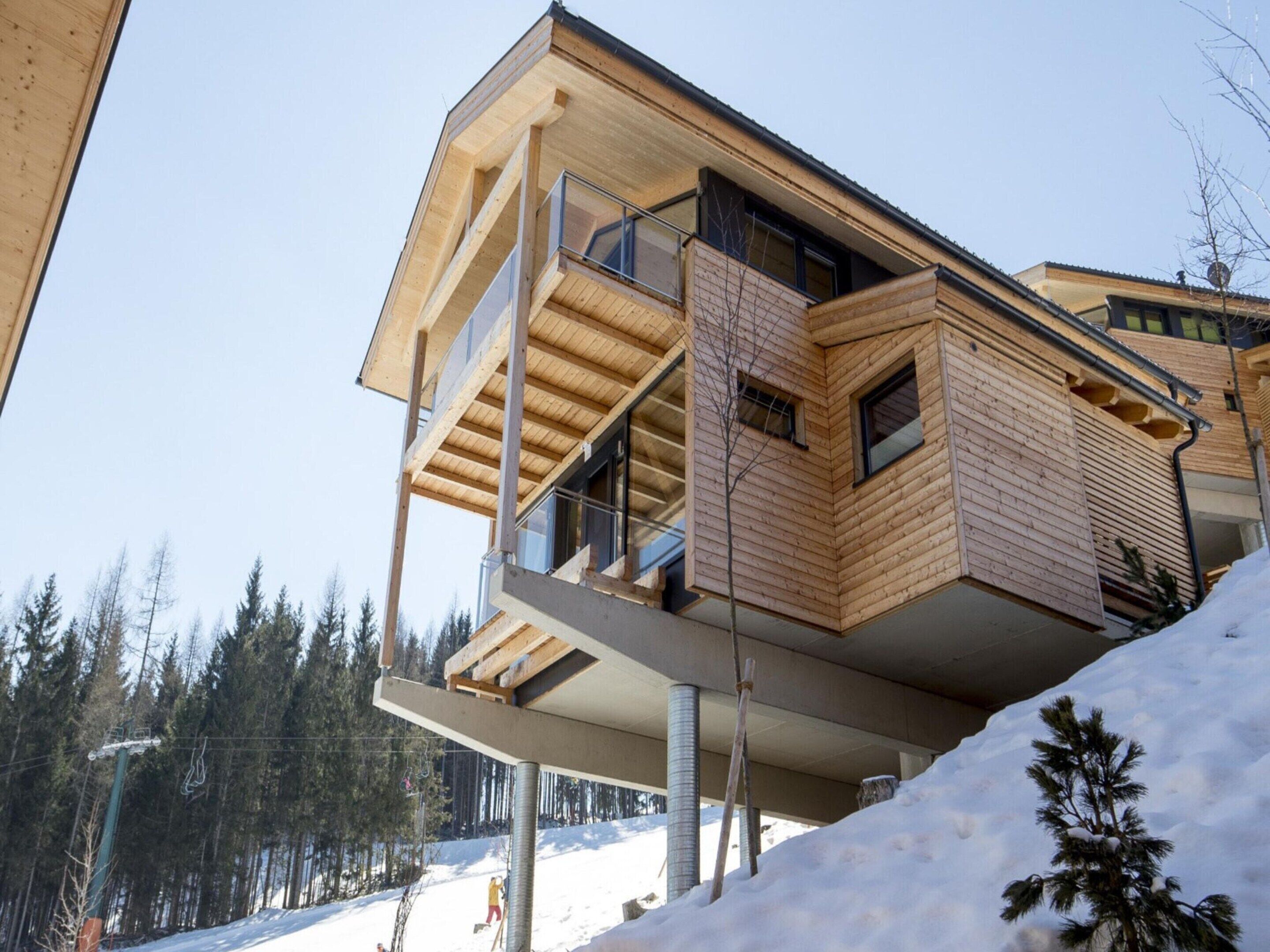 A modern wooden building with a balcony, situated on a snowy slope with a ski lift in the background.
