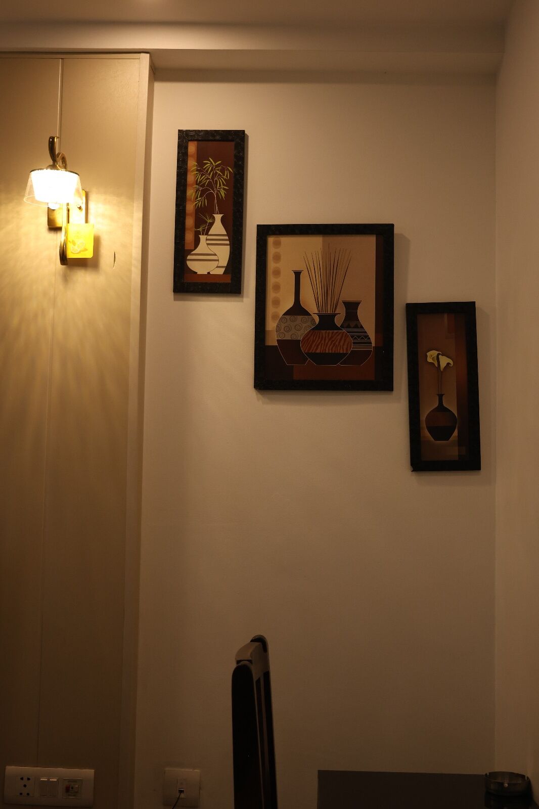Ein Zimmer mit einer Wandlampe, drei gerahmten Kunstwerken mit Vasenmotiven und einem Fernseher.