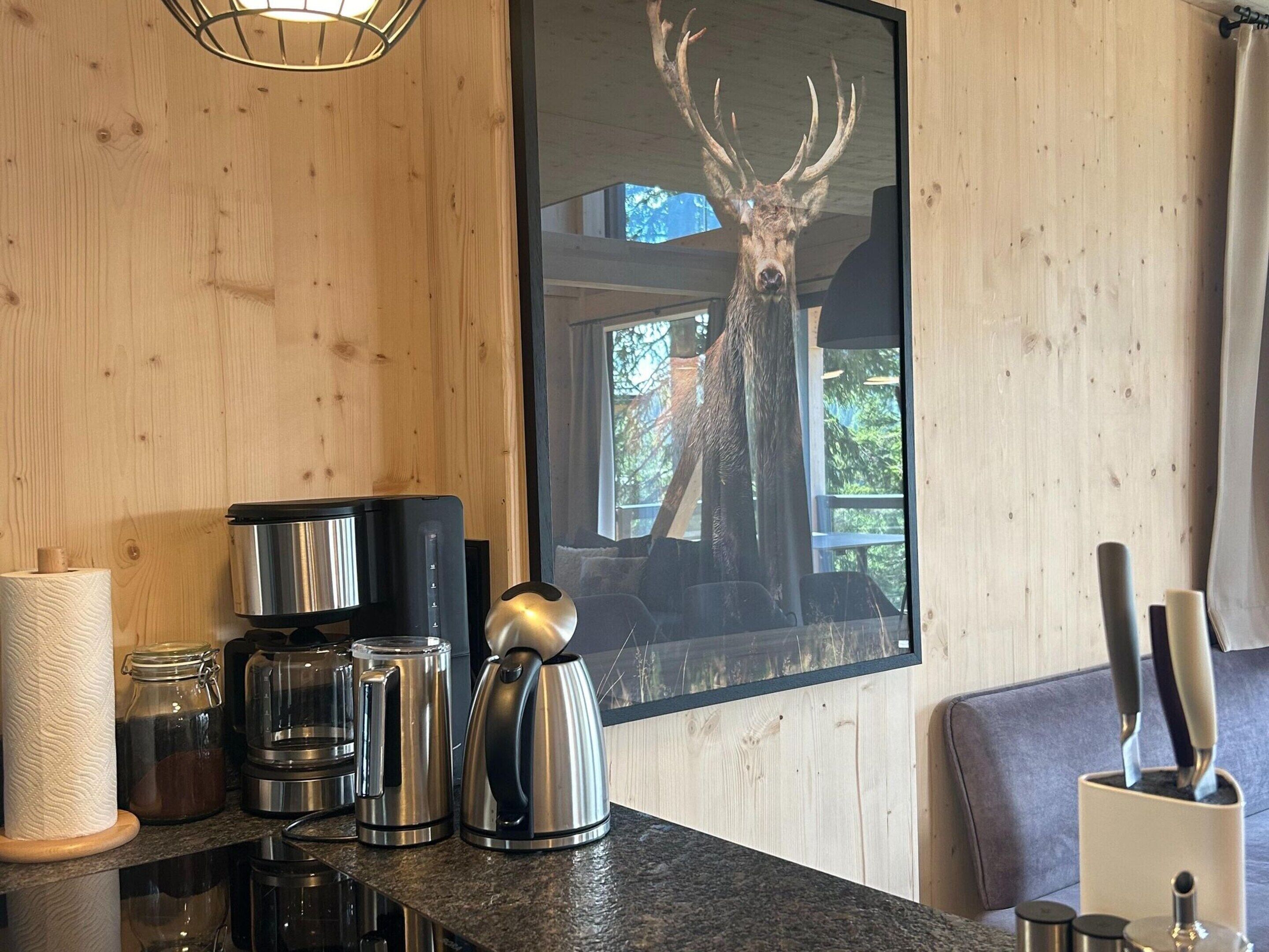En köksbänk med rostfria köksmaskiner, en väggpanel i trä och en inramad tavla med ett rådjurshuvud.