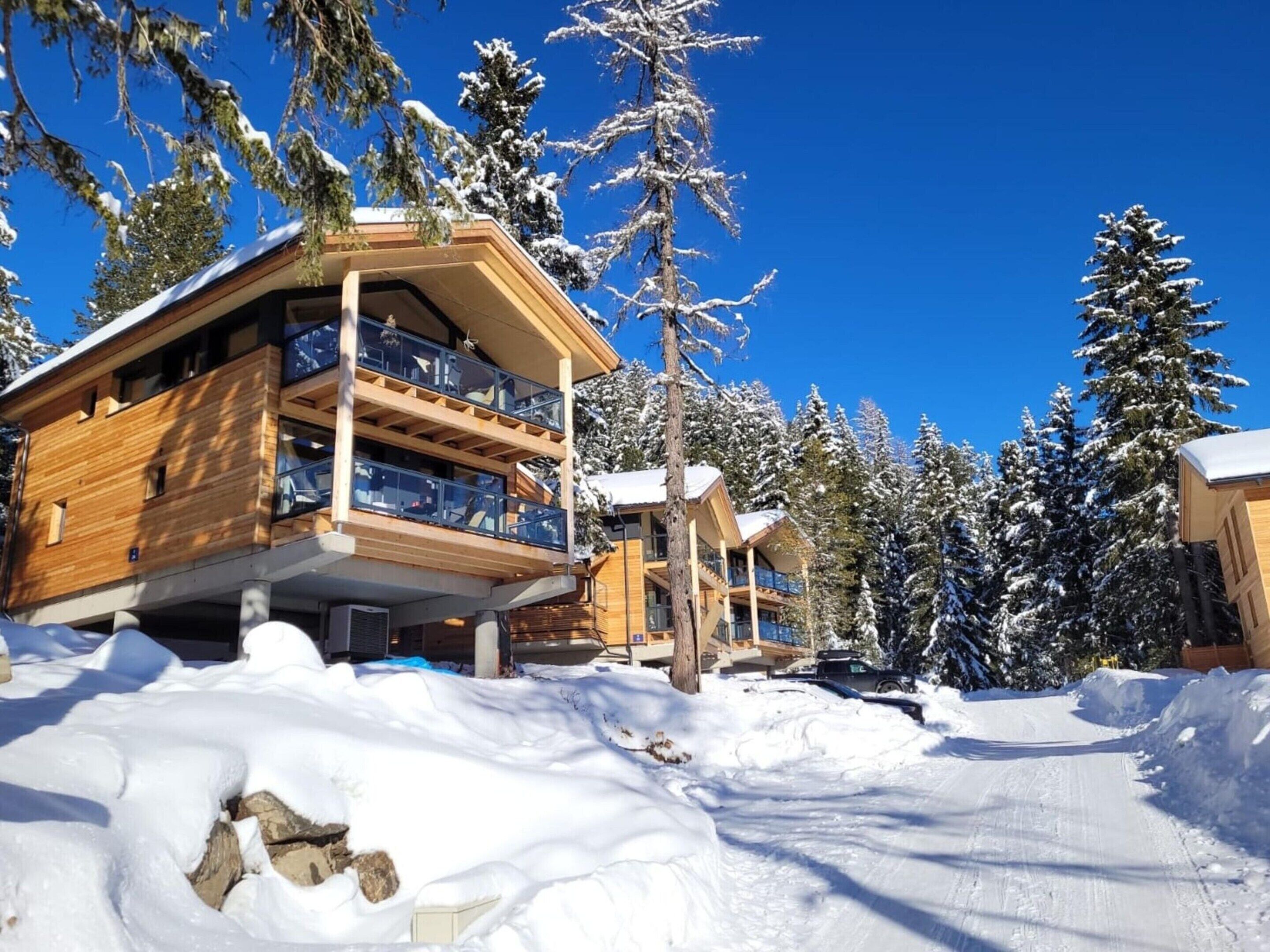A wooden chalet with a balcony, surrounded by snow-covered trees and a clear blue sky.